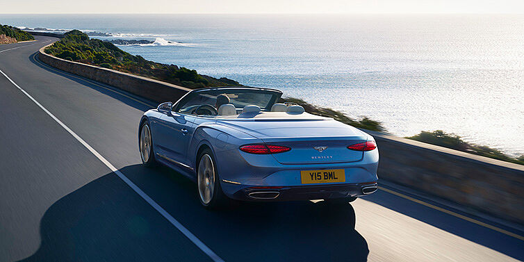 Bentley Kyiv New Bentley Continental GTC Azure convertible rear view in Light Windsor blue paint with 22 inch ten spoke wheels driving dynamically on an ocean road in the sunshine